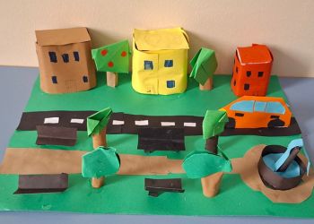 Powiększ zdjęcie: Projekt miasta przedstawiający domy, drzewa, ulicę z jadącym samochodem, park z drzewami, ławkami i fontanną.