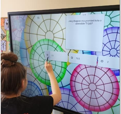 Zadanie na monitorze interaktywnym - Jaką długość ma promień koła o obwodzie PI? Spośród odpowiedzi 1/2, PI/2 i 2 uczennica zaznacza 1/2.